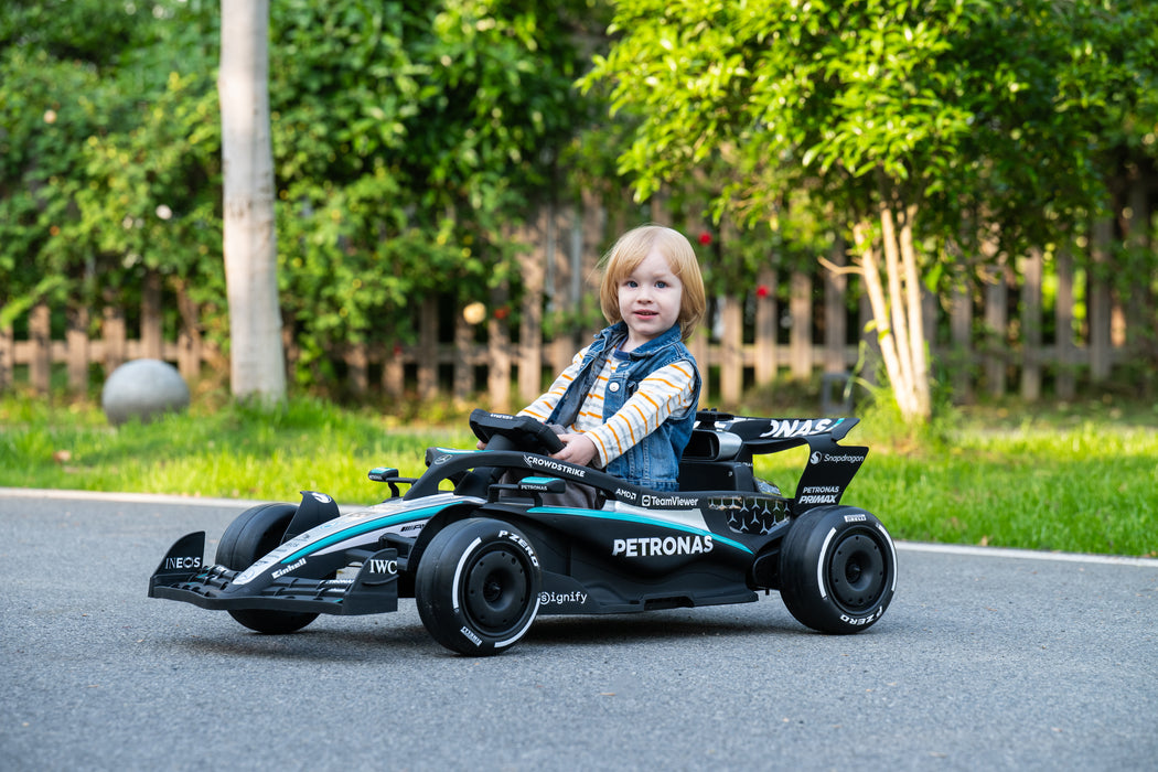 A young child sitting in a black and teal Petronas Formula 1 toy race car on an asphalt path outdoors with trees and a fence in the background. Mercedes F1 W16 ride-on car with parental remote control, two speeds and 50-minute play time
